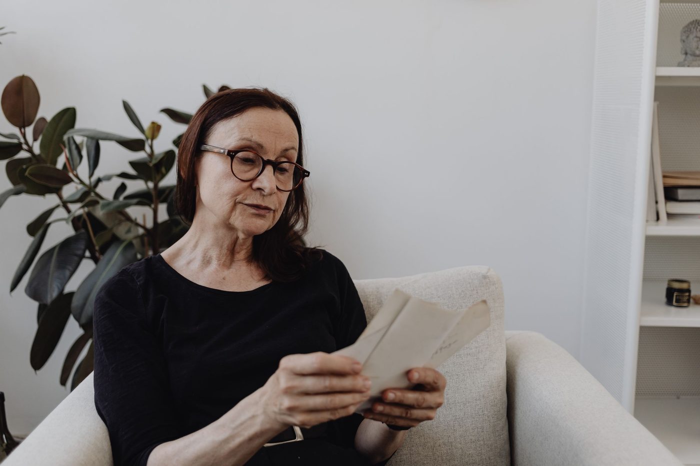 An older woman in glasses sits in a pale armchair reading a letter beside a rubber plant