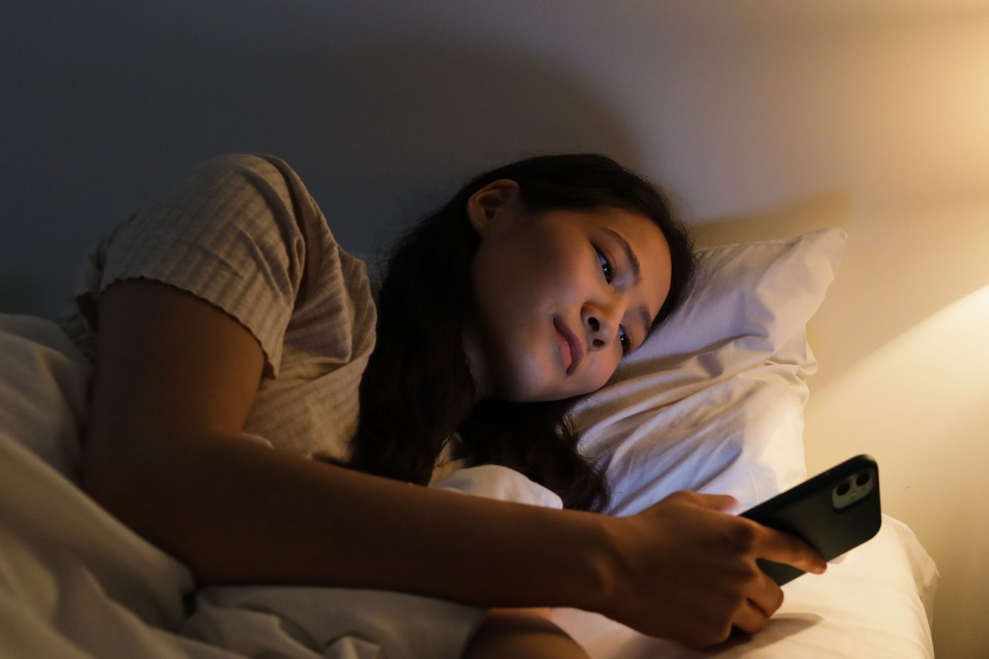 A woman lies in bed under warm bedside light, looking at her phone