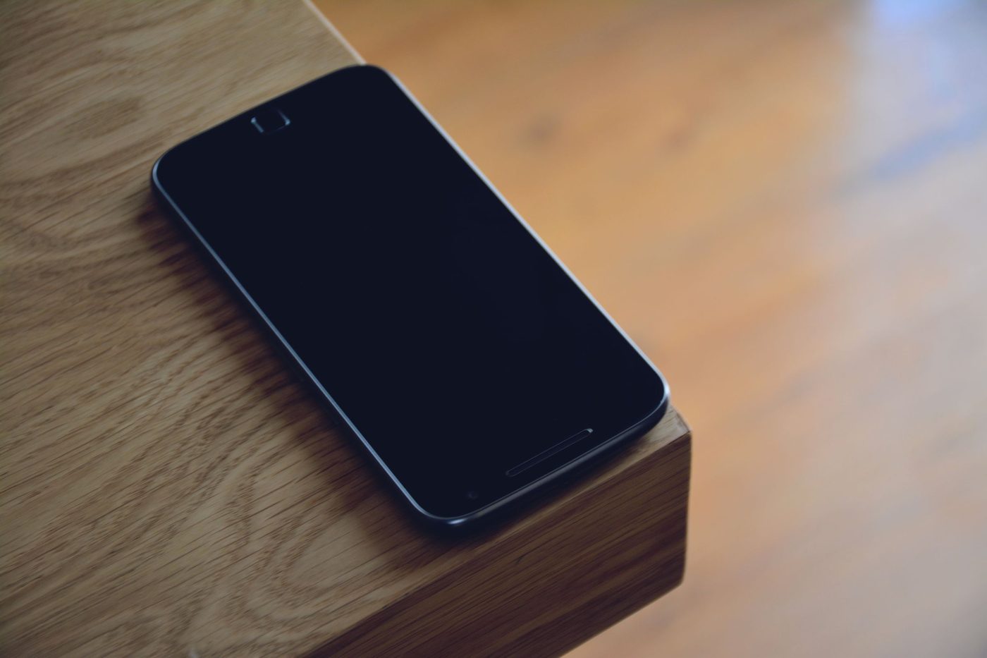 A dark smartphone lies face up on the edge of a light wooden table