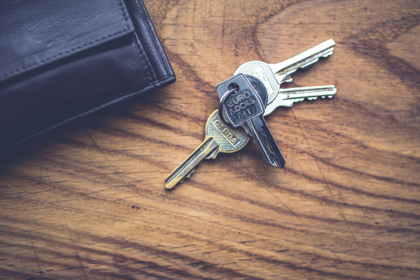 A small bunch of keys lies on a wooden table beside the corner of a black leather wallet