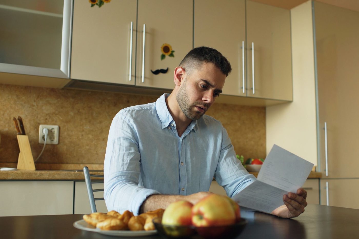 A man in a pale blue shirt reads a letter at a kitchen table with a bowl of apples in front of him