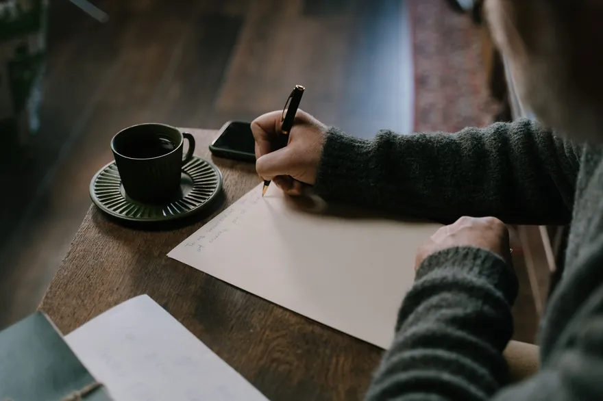 A hand writing on a sheet of paper at a wooden table, a dark cup on a saucer beside it