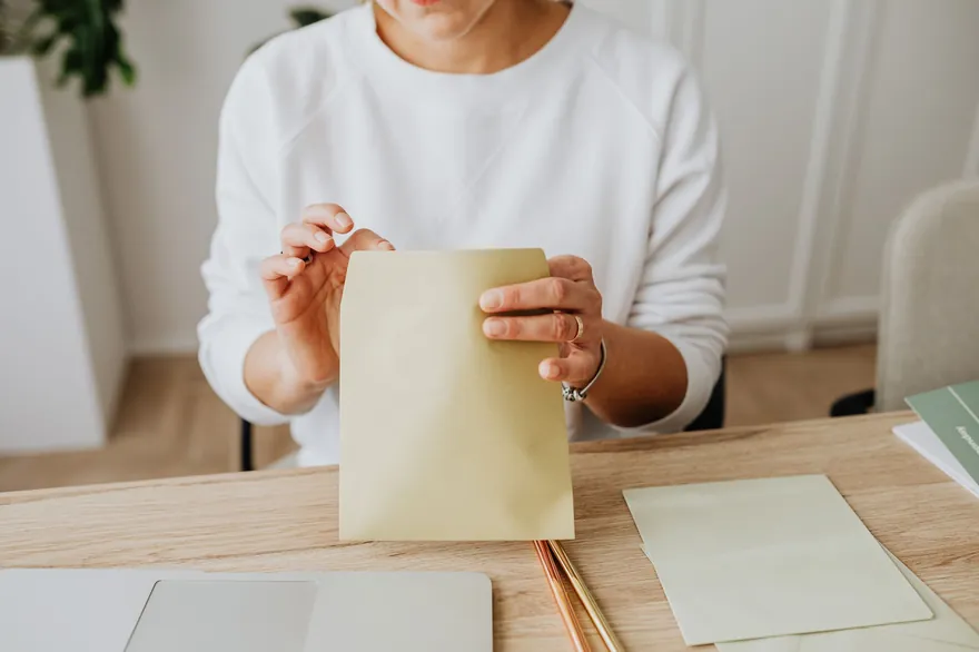 A woman in a white jumper sealing a pale envelope at a light wooden desk