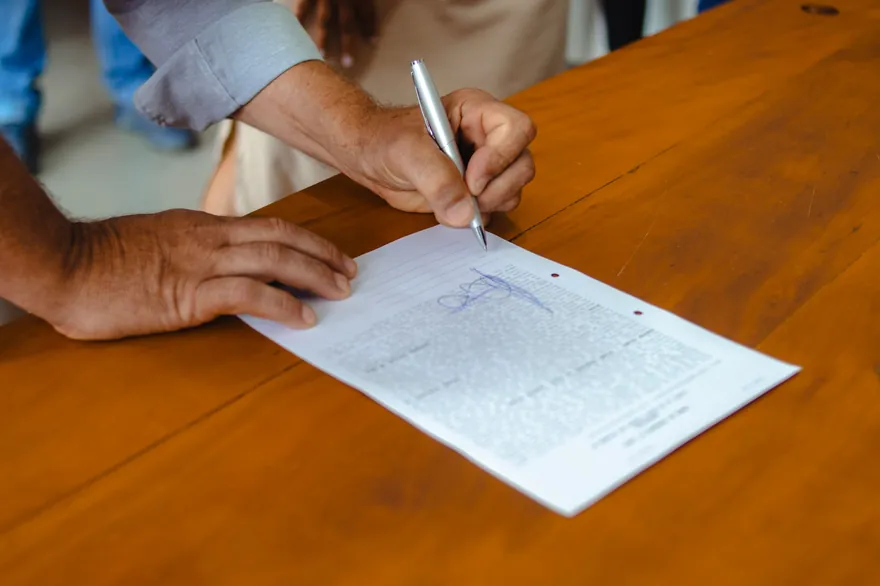 Two hands signing a printed document with a silver pen on a wooden table