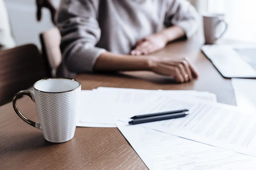 A mug, two pens and printed papers on a dark wooden table, with a person’s arms resting on it