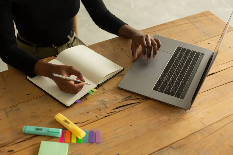 A person&rsquo;s hands rest on an open notebook beside a laptop and highlighters on a wooden table