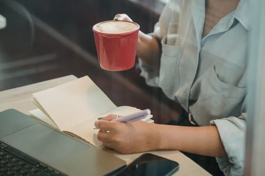 A hand writes in a notebook beside a red mug and a laptop