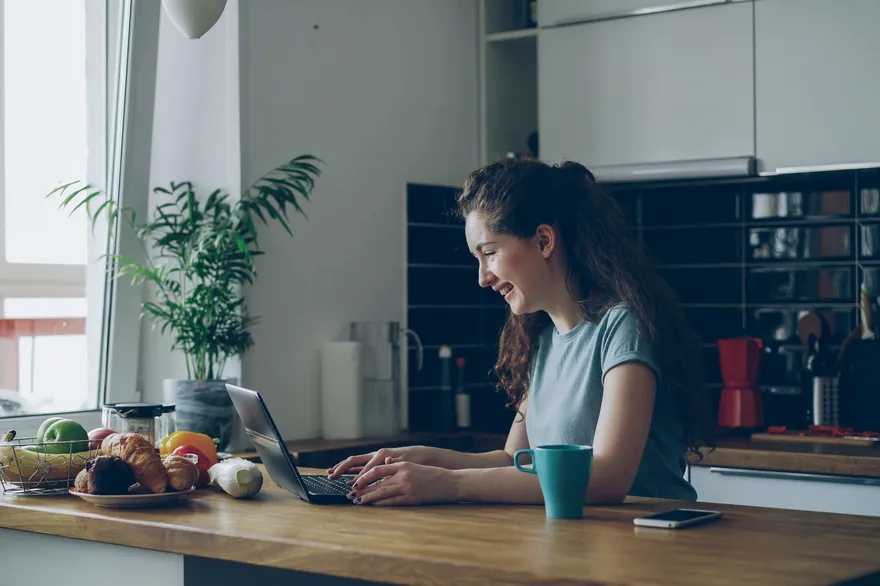 A woman works on a laptop at a kitchen counter with a cup and a bowl of fruit