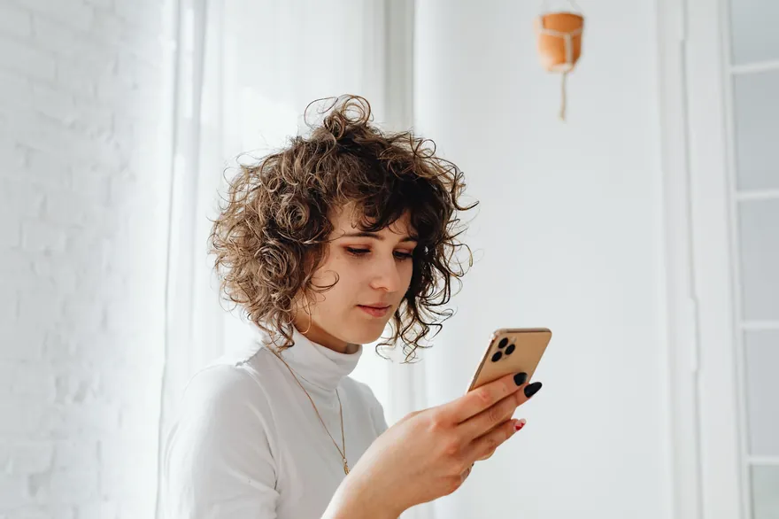 A woman with curly hair stands by a white wall and looks at her phone