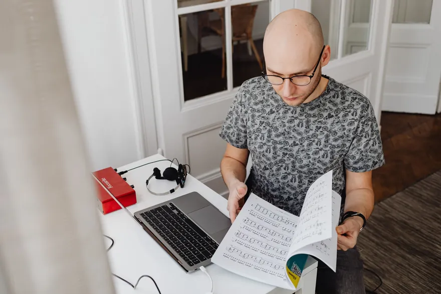 A man sits at a white desk beside a laptop, reading a stack of printed pages