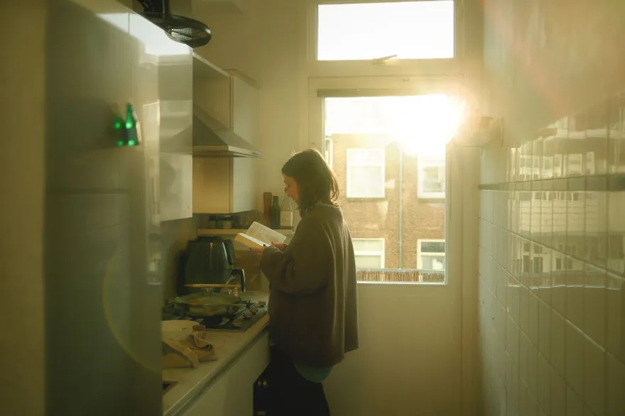 A woman in a loose cardigan reading a book in a narrow kitchen, late sunlight coming through the window behind her