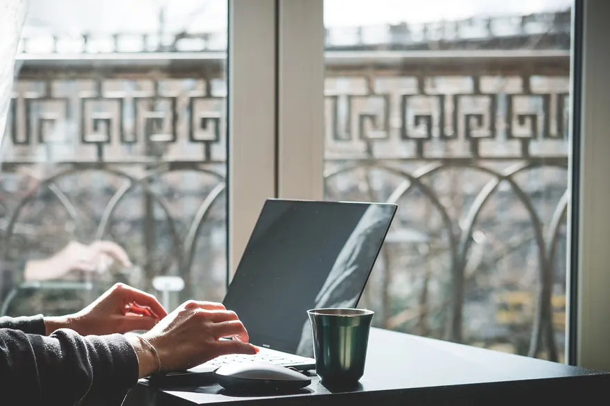 Hands typing on a laptop at a desk beside a window with a balcony railing and a green mug