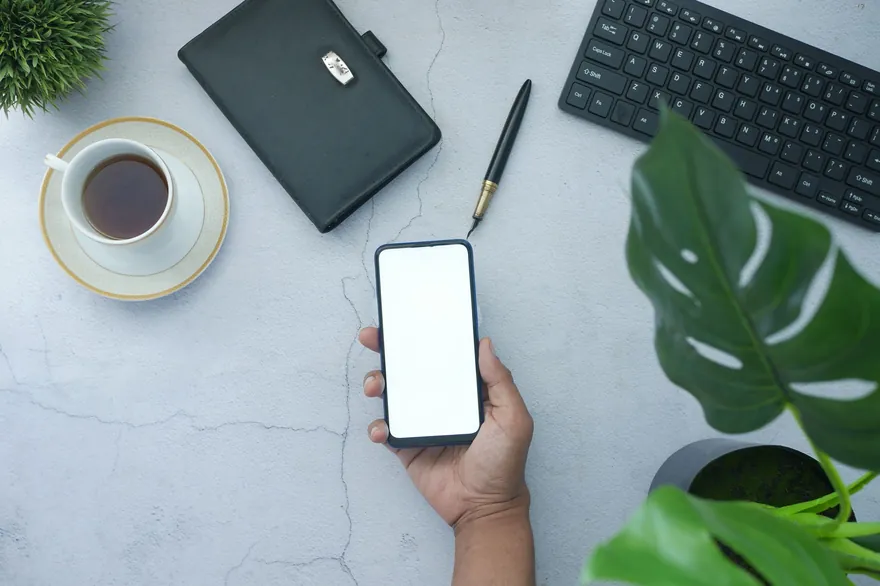 A hand holds a phone with a blank screen above a desk with a coffee cup, a black folder and a pen