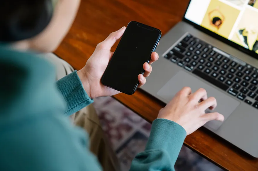 A person holds a phone above an open laptop on a wooden desk