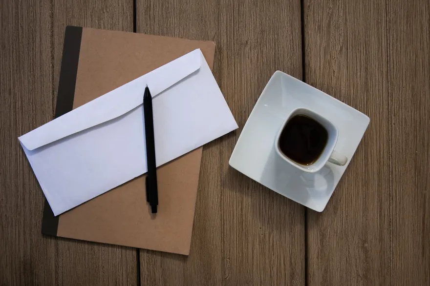 A white envelope and a pen on a brown notebook beside a cup of coffee on a wooden table
