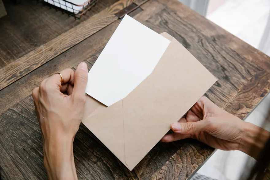 Hands opening an envelope with a folded card on a dark wooden desk, seen from above