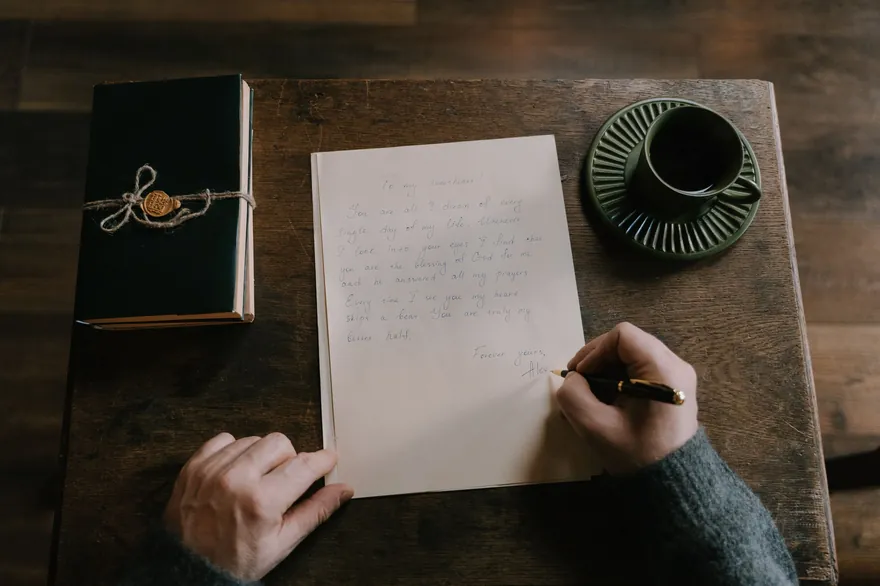A hand writes a letter on a wooden table beside a closed journal and a coffee cup