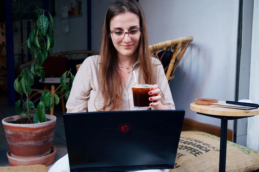 A woman sits with a drink in a cafe, her laptop open in front of her