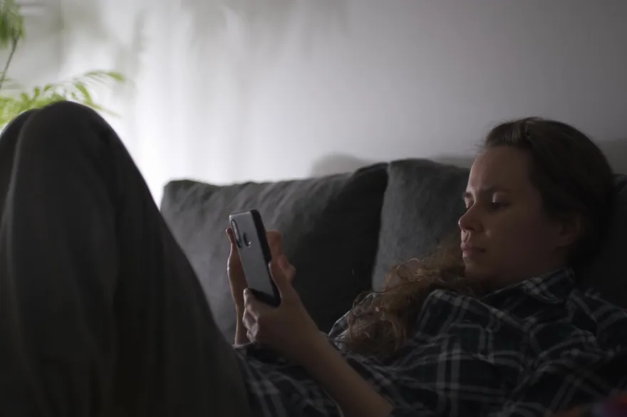 A woman sits on a sofa in a dim room, her face lit as she looks at her smartphone