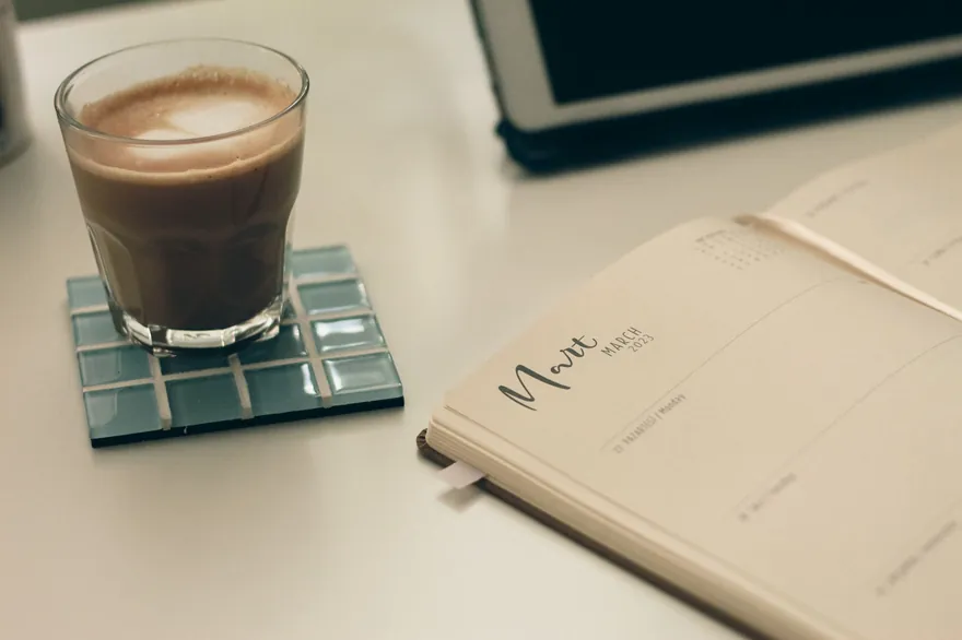 An open diary beside a glass of coffee on a glass coaster