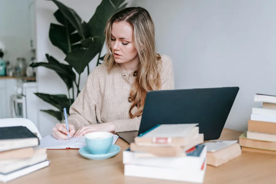 A woman with long fair hair writes in a notebook at a table beside a laptop, a cup of tea and a pile of books