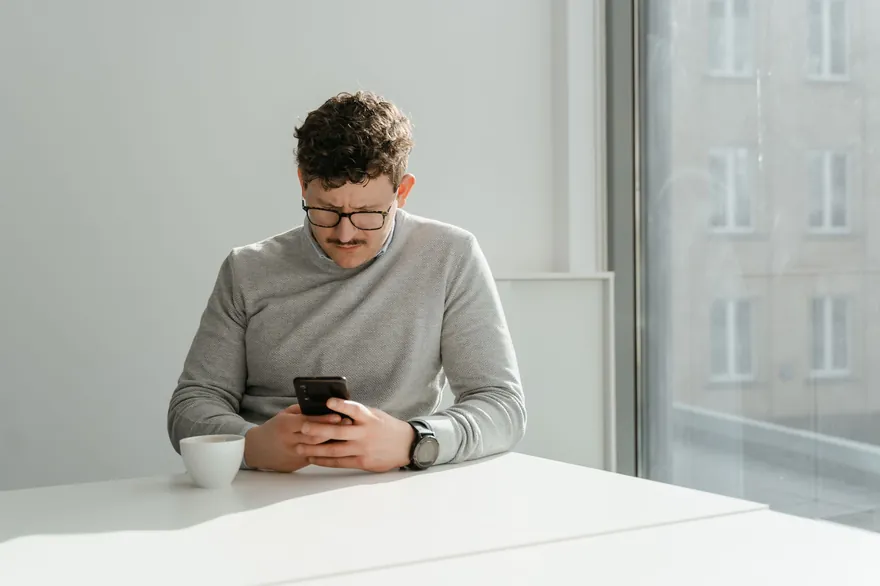 A man in glasses and a grey jumper frowning at the phone in his hands at a white table by a window