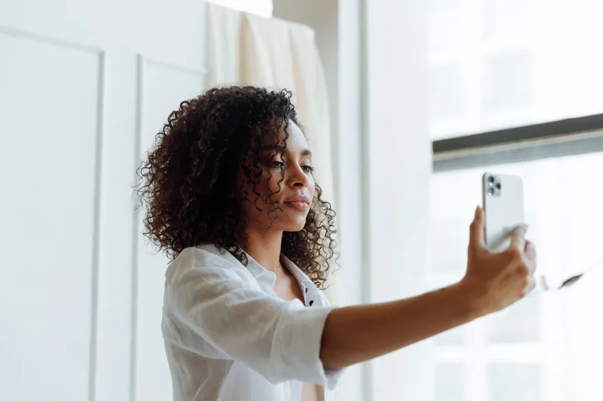 A woman with curly hair by a bright window holding a phone at arm&rsquo;s length to film a selfie, the back of the phone towards the camera