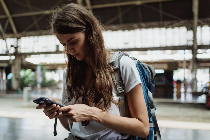 A woman with a blue backpack looking down at her phone on a covered railway platform