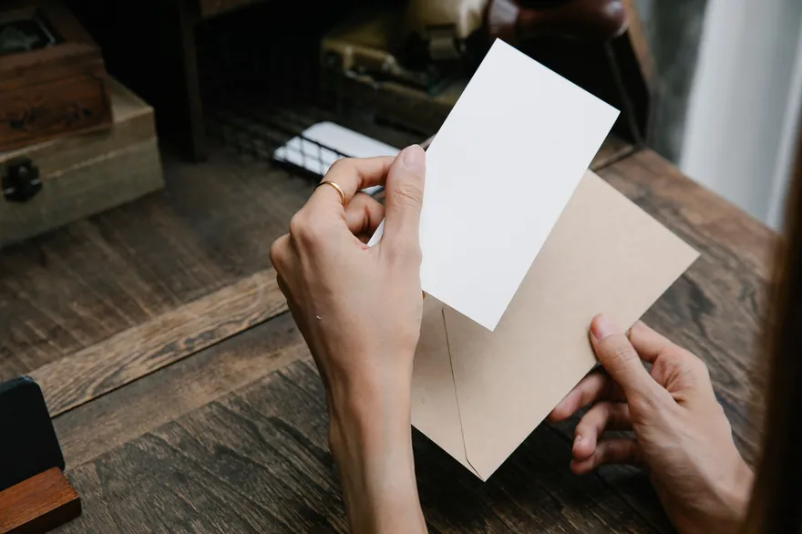 Two hands hold a blank white card above a kraft envelope on a wooden desk