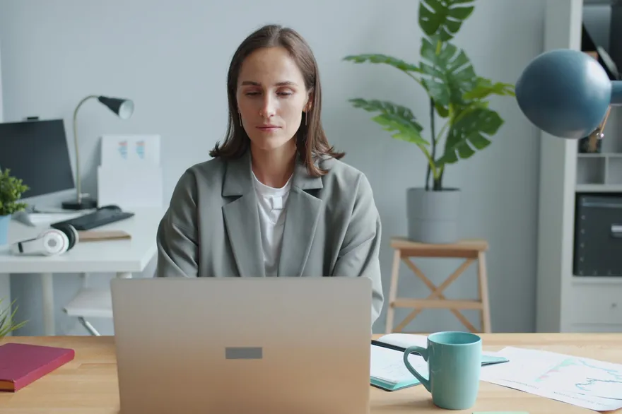 A woman sits at a desk in a bright office, looking at her laptop with a notebook beside it