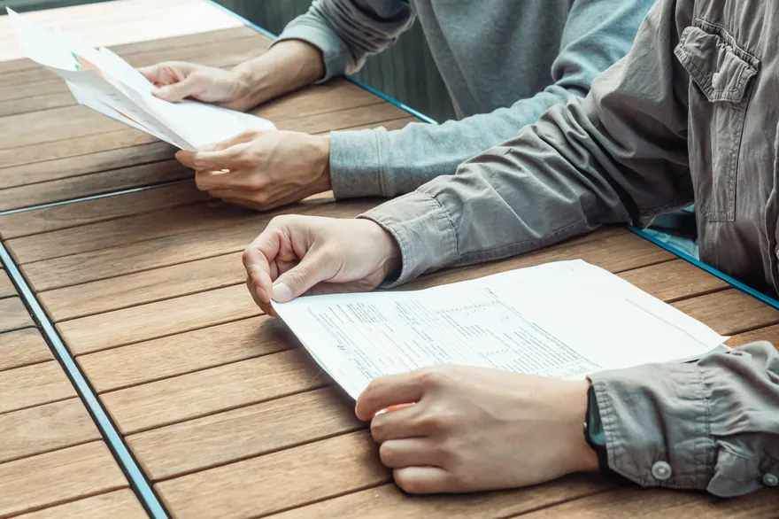 Two people sit at a wooden slatted table, one holding a printed form